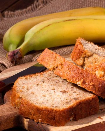 Banana bread on a wood cutting board and 2 bananas in the background