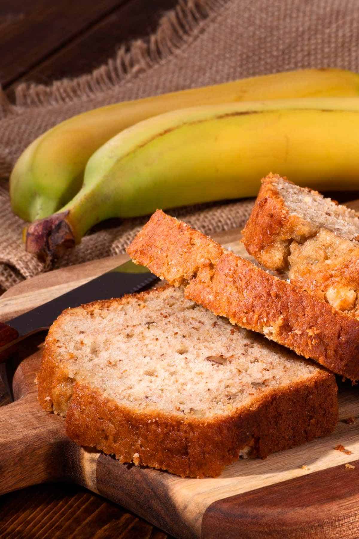 Banana bread on a wood cutting board and 2 bananas in the background